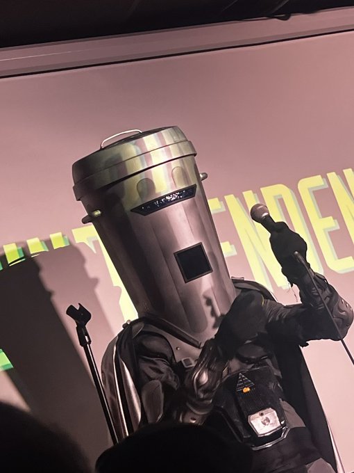 Count Binface — a polished metal bin for a helmet, black space armour — holding a microphone on stage